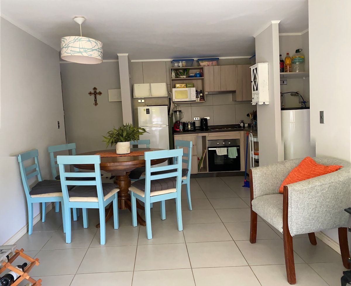 An open-plan dining area features a round wooden table surrounded by six light blue chairs. A kitchen is visible in the background, equipped with a refrigerator, stove, and microwave. A cozy seating arrangement is present, consisting of a grey armchair with an orange accent pillow.