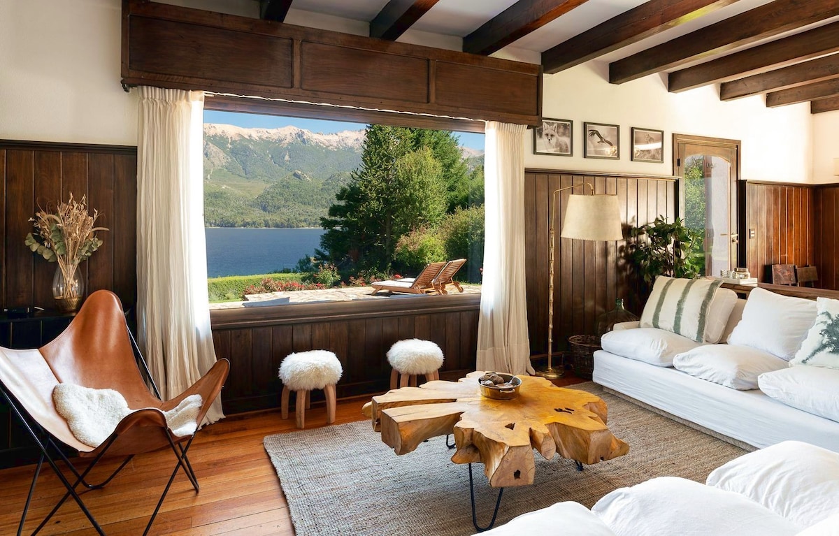 A cozy living area features wooden beams and warm oak floors. Large windows frame views of the lake and distant mountains. A unique wooden coffee table sits at the center, surrounded by a plush sectional and a leather chair. Soft, decorative elements enhance the space.