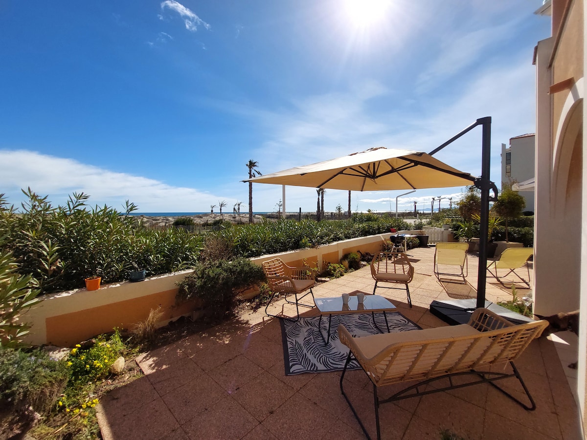 An outdoor terrace features a sunshade canopy providing shade over comfortable seating. A low table sits in the center, surrounded by lounge chairs. Lush green plants frame the space, and a view of the ocean is visible in the background under a clear blue sky.
