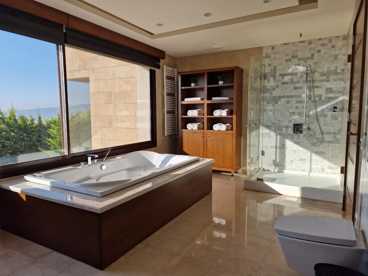 A spacious bathroom is highlighted by a large soaking tub next to a window with a view. A modern shower stall with glass doors is located in the corner, while warm wooden shelving displays neatly rolled towels. Natural light enhances the calming atmosphere.
