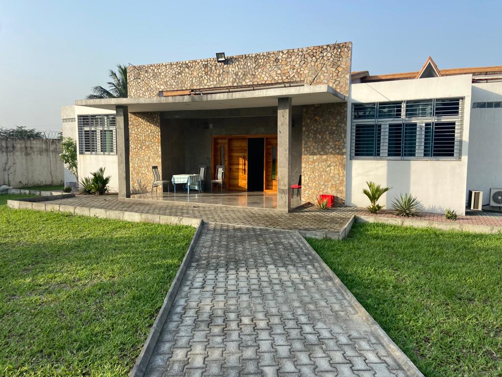 The exterior of a modern house features a stone-facade entrance and a spacious front porch with seating. A neatly paved walkway leads to the entrance, surrounded by green grass and small decorative plants. Large windows reflect sunlight, enhancing the inviting ambiance.