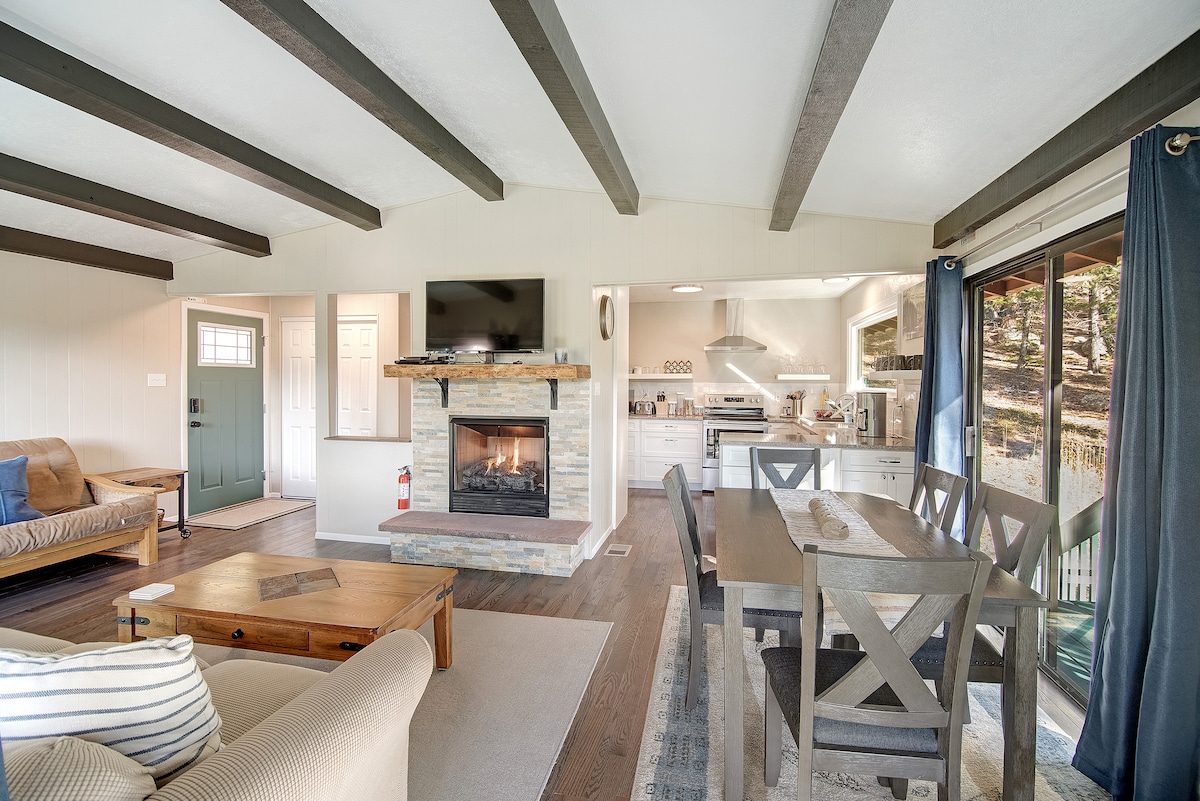 A spacious living area features exposed wooden beams and a cozy fireplace framed by stone. A dining table with six chairs is positioned nearby, and a well-equipped kitchen is visible in the background. Large windows allow natural light and provide views of the surrounding outdoors.