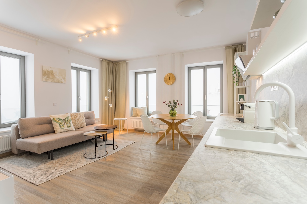 A spacious living area features large windows allowing natural light to fill the room. A comfortable sofa sits alongside a round dining table with four chairs. A modern kitchenette, highlighted by sleek countertops, provides essential appliances. Decorative elements create a welcoming atmosphere.