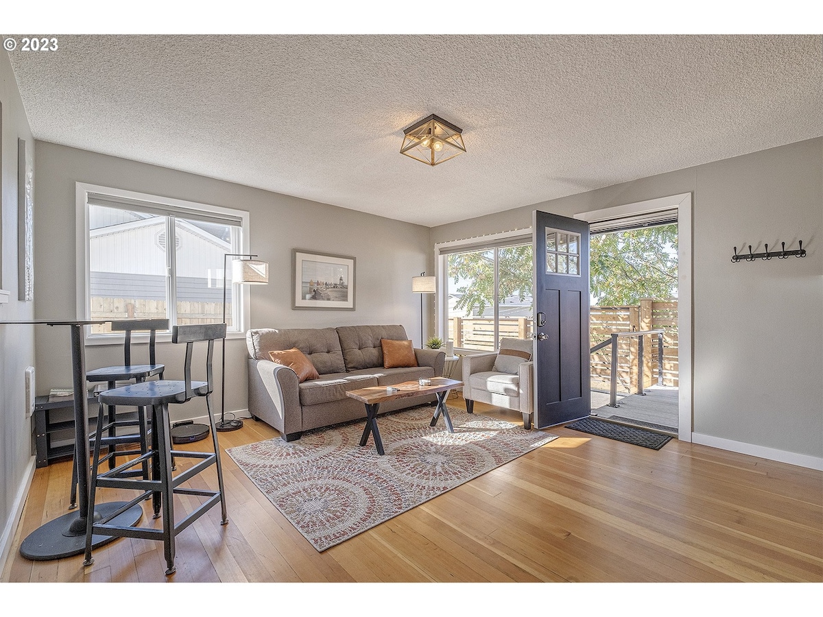 A bright living area features a comfortable gray couch and an armchair, with natural light filtering through large windows. A coffee table sits on a patterned rug, while a small dining set is positioned nearby. The front door opens to a deck, enhancing indoor-outdoor flow.