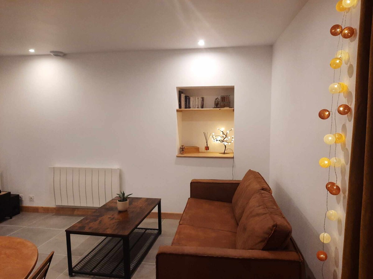 A simple living area features a brown couch positioned next to a wooden coffee table. A small shelf is built into the wall, displaying books and decorative items. Soft lighting is provided by recessed ceiling lights and a string of decorative lights along one corner.