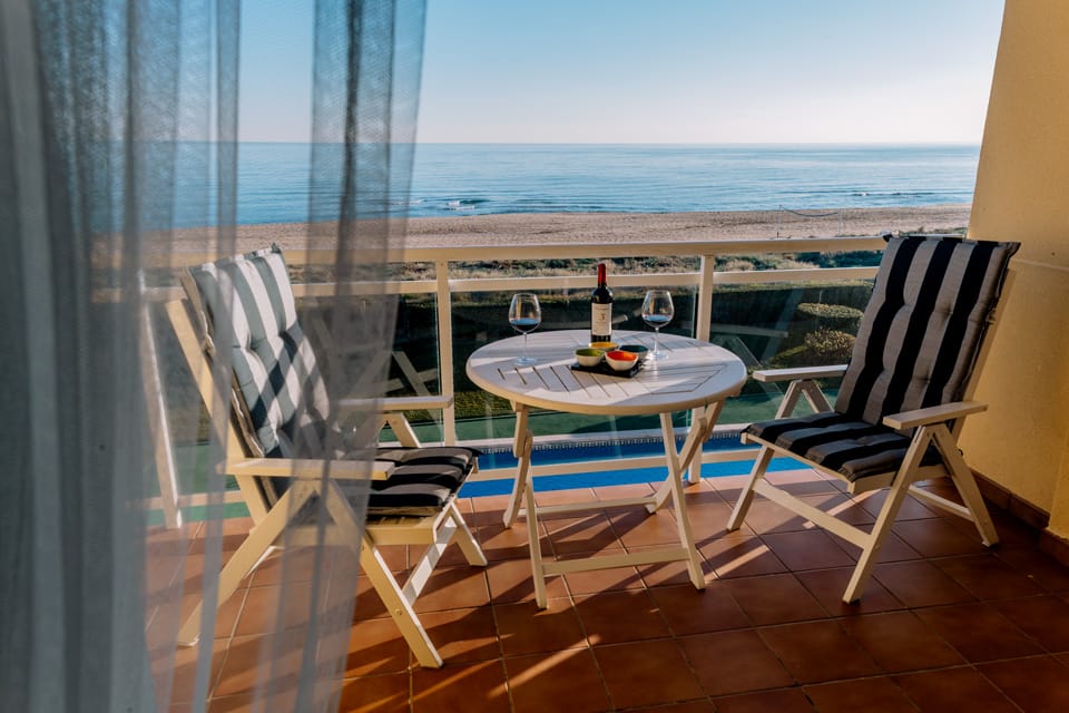 A small balcony features a round table with striped chairs overlooking the beach. A bottle of wine and two glasses are set on the table, with the calm sea visible in the background, providing a serene view.