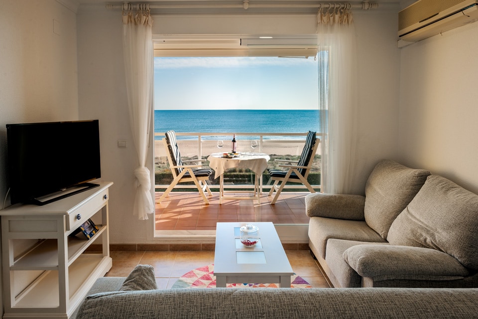 A living area features a comfortable gray sofa with colorful cushions, overlooking a terrace. A glass door opens to a beachfront view, revealing a table set with two chairs and a bottle. A flat-screen TV is positioned on a white cabinet.