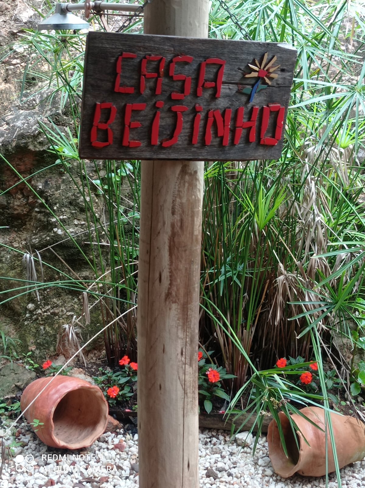A wooden sign displaying the name 'Casa Beijinho' in bright red letters is prominently positioned. Surrounding greenery adds a natural touch, while terracotta pots are placed decoratively on a gravel surface adorned with small red flowers.