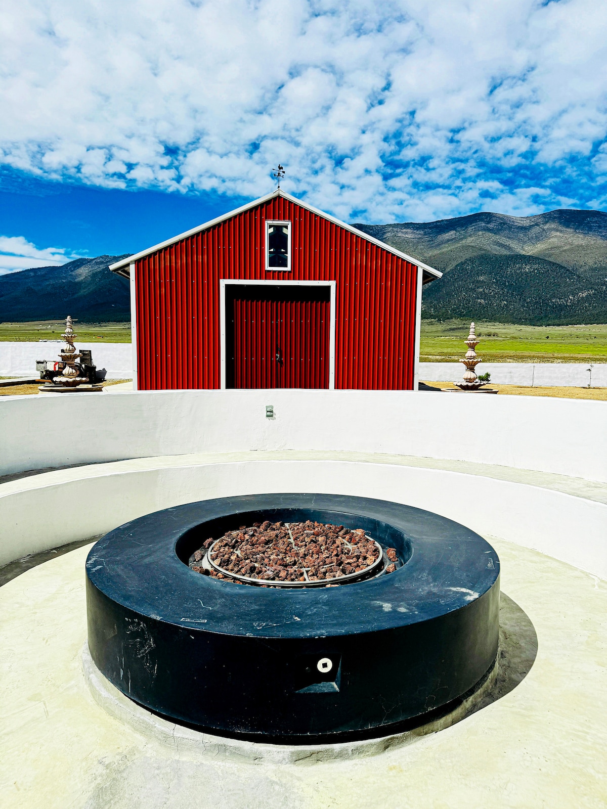 A circular fire pit is set in the foreground, surrounded by a smooth, white concrete structure. Behind it, a bright red barn stands against a backdrop of rolling green hills under a partly cloudy sky.