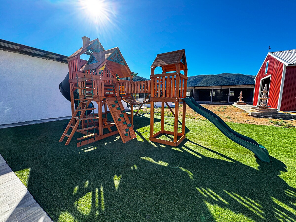 A wooden playground structure is situated on a grassy area, featuring slides, climbing walls, and playhouses. Sunlight casts shadows on the equipment, with a red barn visible in the background under a clear blue sky.