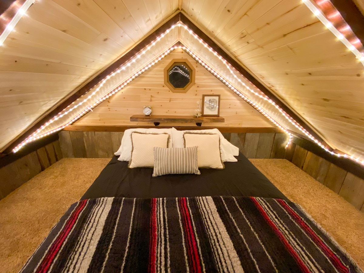A cozy sleeping area is positioned under a vaulted wooden ceiling adorned with soft lighting. The bed features a dark comforter accented by decorative pillows and is complemented by a patterned blanket. Natural wood elements and simple decor enhance the inviting space.