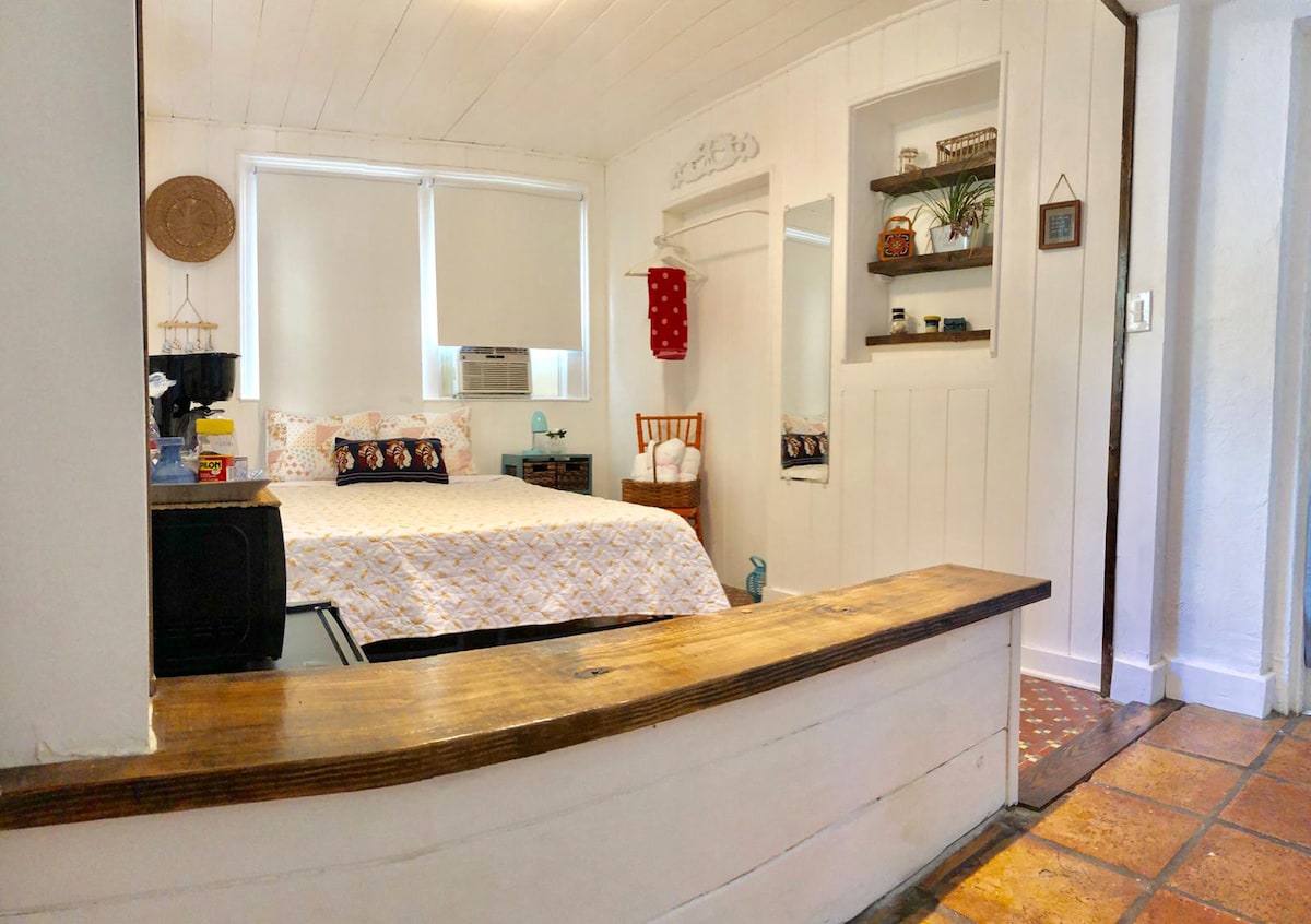 A cozy bedroom is designed with a white shiplap wall, featuring a queen bed covered in patterned bedding. An air conditioning unit is positioned near a window, with natural light illuminating the space. A decorative shelving unit displays plants and decor, complementing the inviting atmosphere.