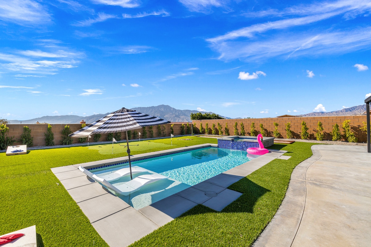 An inviting pool area is surrounded by lush artificial turf and vibrant desert landscaping. A sun umbrella provides shade over the pool's ledge lounge chairs, while a pink inflatable floatie adds a touch of fun. Mountain views stretch across the horizon under a clear blue sky.