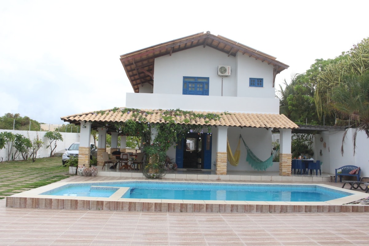 A two-story villa is depicted with a spacious outdoor area featuring a rectangular swimming pool. A covered patio is present, adorned with hammocks and seating arrangements. Lush greenery enhances the surroundings, while the roof exhibits a traditional design with wooden beams.