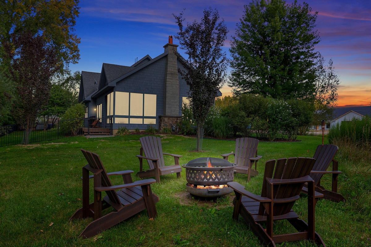 A serene outdoor space features a circular arrangement of dark wooden Adirondack chairs surrounding a fire pit. The lawn is green and expansive, with trees and shrubbery providing a natural backdrop. A charming home with large windows is visible in the background, illuminated by sunset hues.