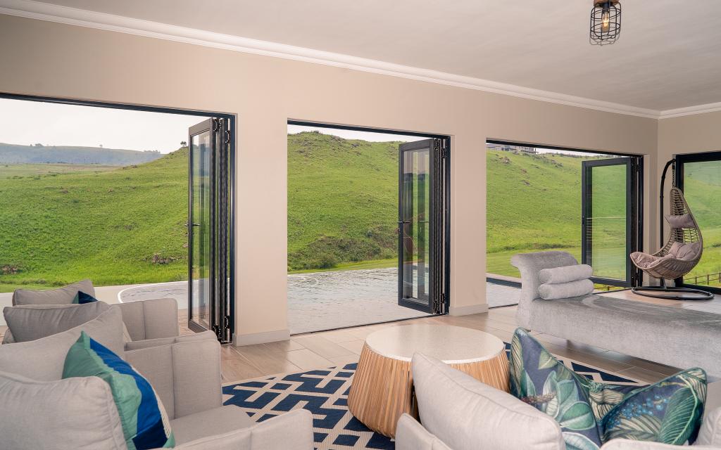 A spacious lounge area features modern furnishings in neutral tones with touches of blue. Large glass doors open to a scenic view of rolling green hills, enhancing natural light. A circular wooden coffee table is centered, complementing a cozy seating arrangement with plush cushions.