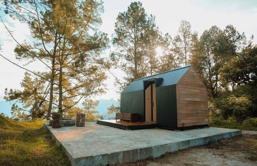A modern cabin structure is set on a concrete platform, surrounded by tall trees. The exterior features a combination of wooden and dark panels, with a sloped roof. Soft sunlight filters through the trees, creating a serene outdoor atmosphere.