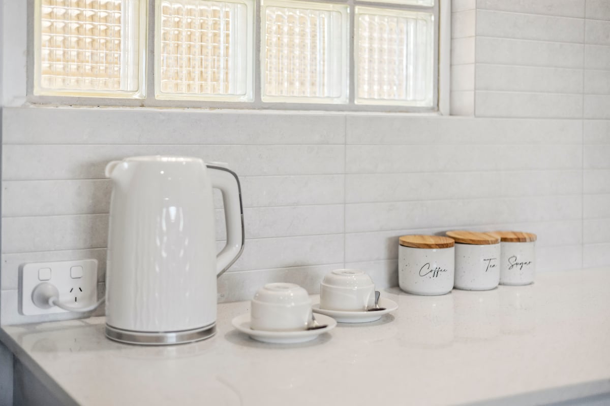 An equipped kitchen corner is displayed with a white electric kettle, two matching cups on saucers, and ceramic containers labeled for coffee and tea, alongside sugar. The light-colored countertop reflects the natural light coming through a glass block window.