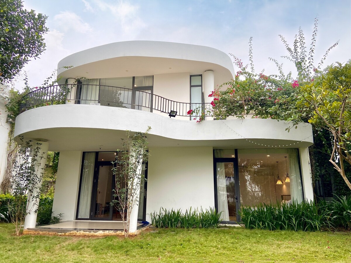 The exterior of the villa showcases a unique architectural design with smooth curves and large glass doors. Lush greenery surrounds the property, complemented by flowering plants. A spacious lawn in front offers a serene outdoor space, perfect for relaxation or play.