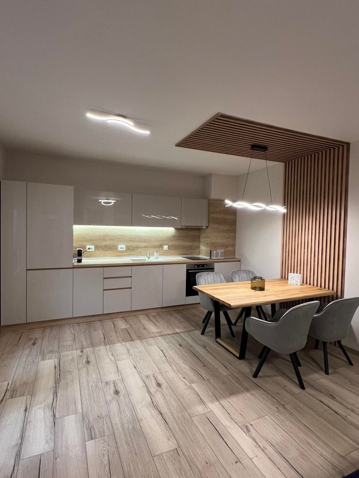 The kitchen area features modern cabinetry with a sleek finish and a wood accent ceiling. A dining table for four is situated beneath a designer light fixture. Essential appliances, including a stove and refrigerator, are neatly integrated, complemented by a warm wood flooring.