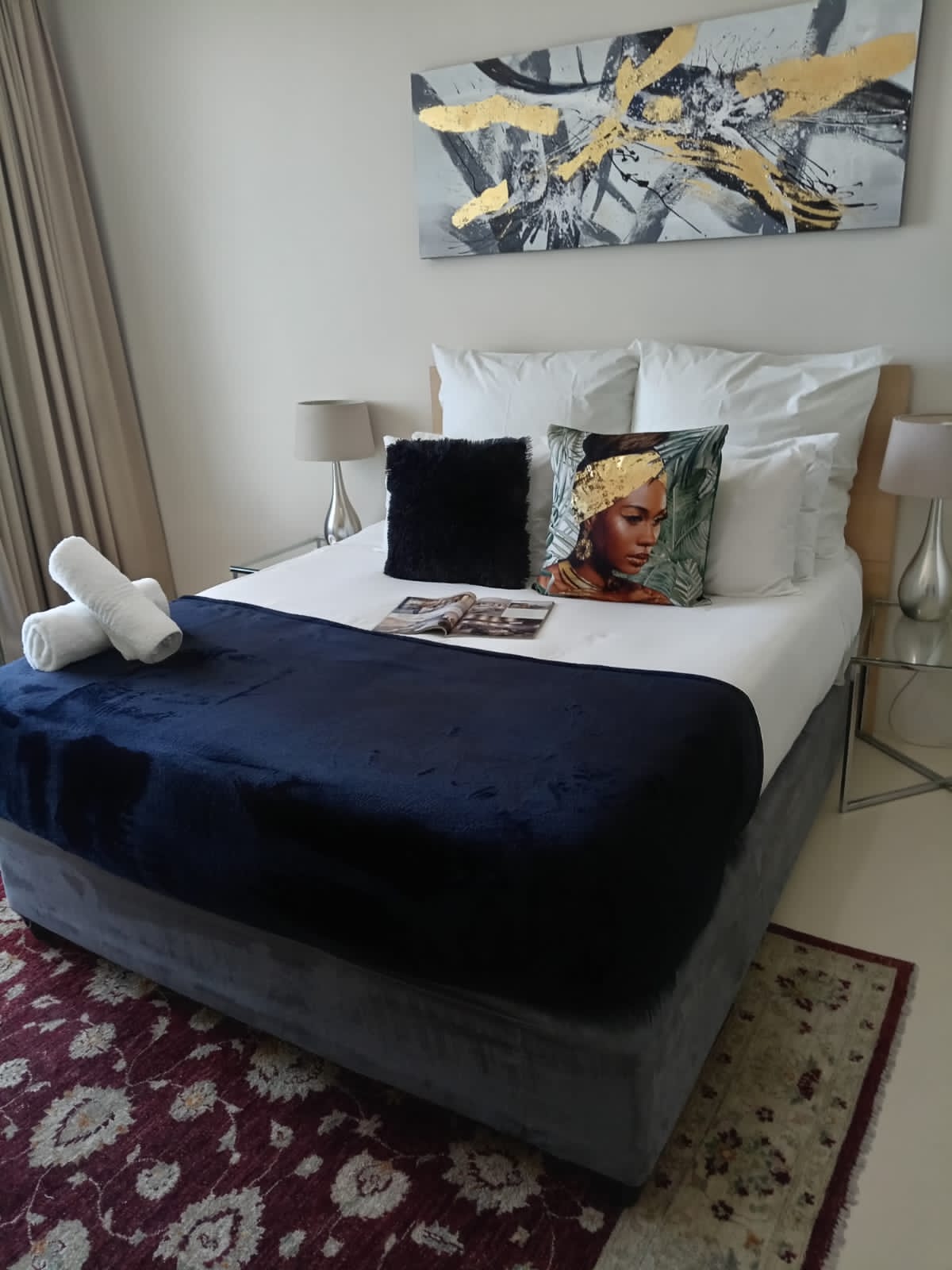 A spacious bed is situated against a light-colored wall, topped with fluffy white pillows and decorative cushions. A dark blue throw covers the bed's lower section. A stylish piece of artwork is mounted above the bed, and two bedside lamps are positioned on either side.