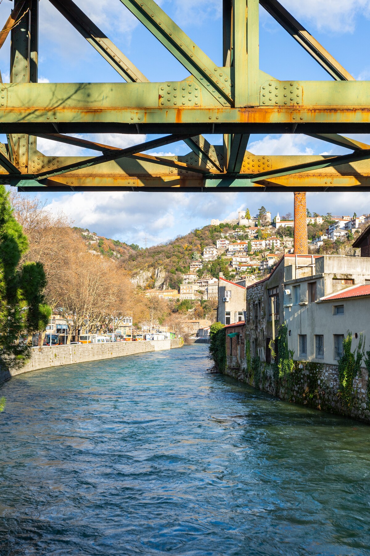 A scenic view is captured through a rusted metal bridge, revealing a flowing river beneath. On the banks, aged buildings show a blend of nature and urban life, while the hillside beyond is adorned with trees and houses, illustrating the harmony of the landscape.