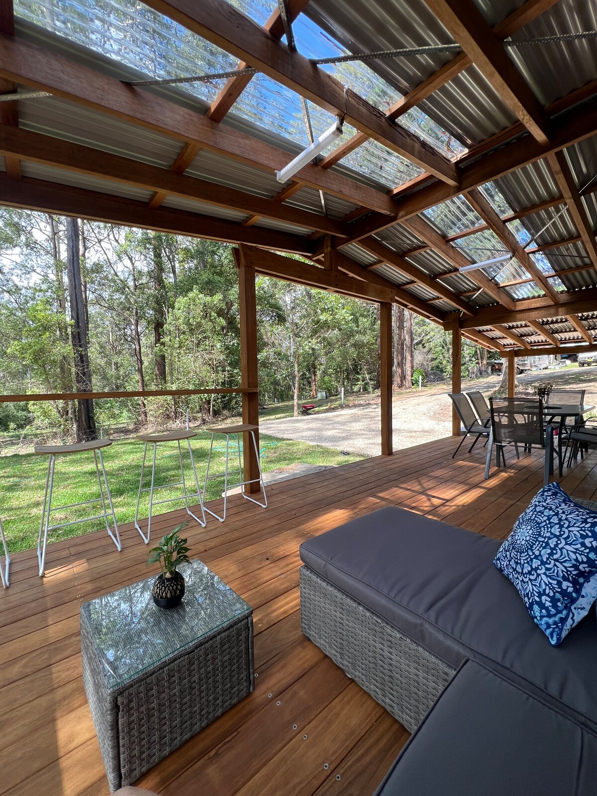 A spacious outdoor deck features a comfortable lounge area with a grey couch adorned with a blue decorative pillow. A glass-topped table is visible, accompanied by metal bar stools facing the green bushland and trees beyond, enhancing the serene setting.