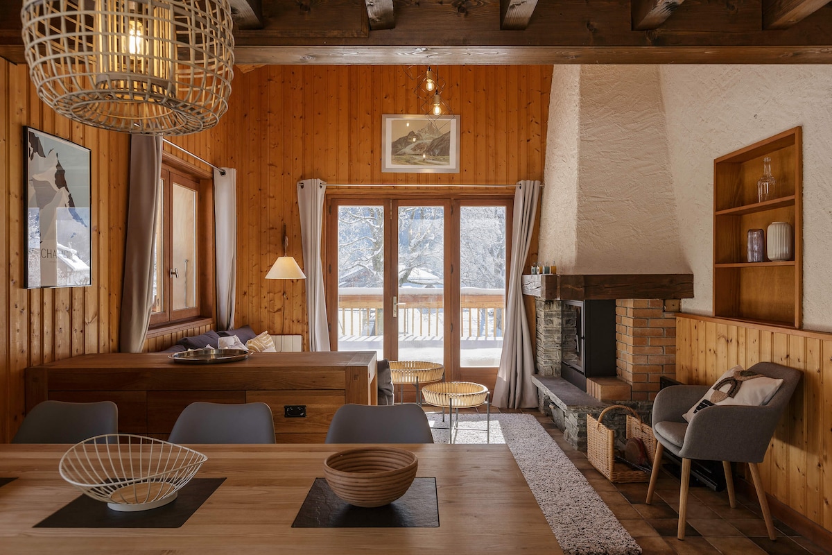 The living area features a wooden ceiling and walls, creating a warm atmosphere. Large glass doors open to the terrace, allowing natural light. A cozy sitting area includes a fireplace, while a dining table is set for gatherings. Modern decor elements are subtly integrated throughout.