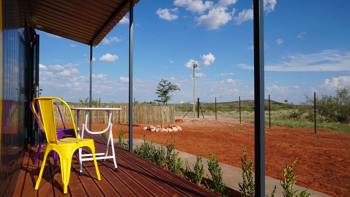 An outdoor patio features two colorful chairs and a small table, inviting relaxation. The surrounding landscape showcases red earth and sparse vegetation under a clear blue sky, with distant fencing marking property boundaries.