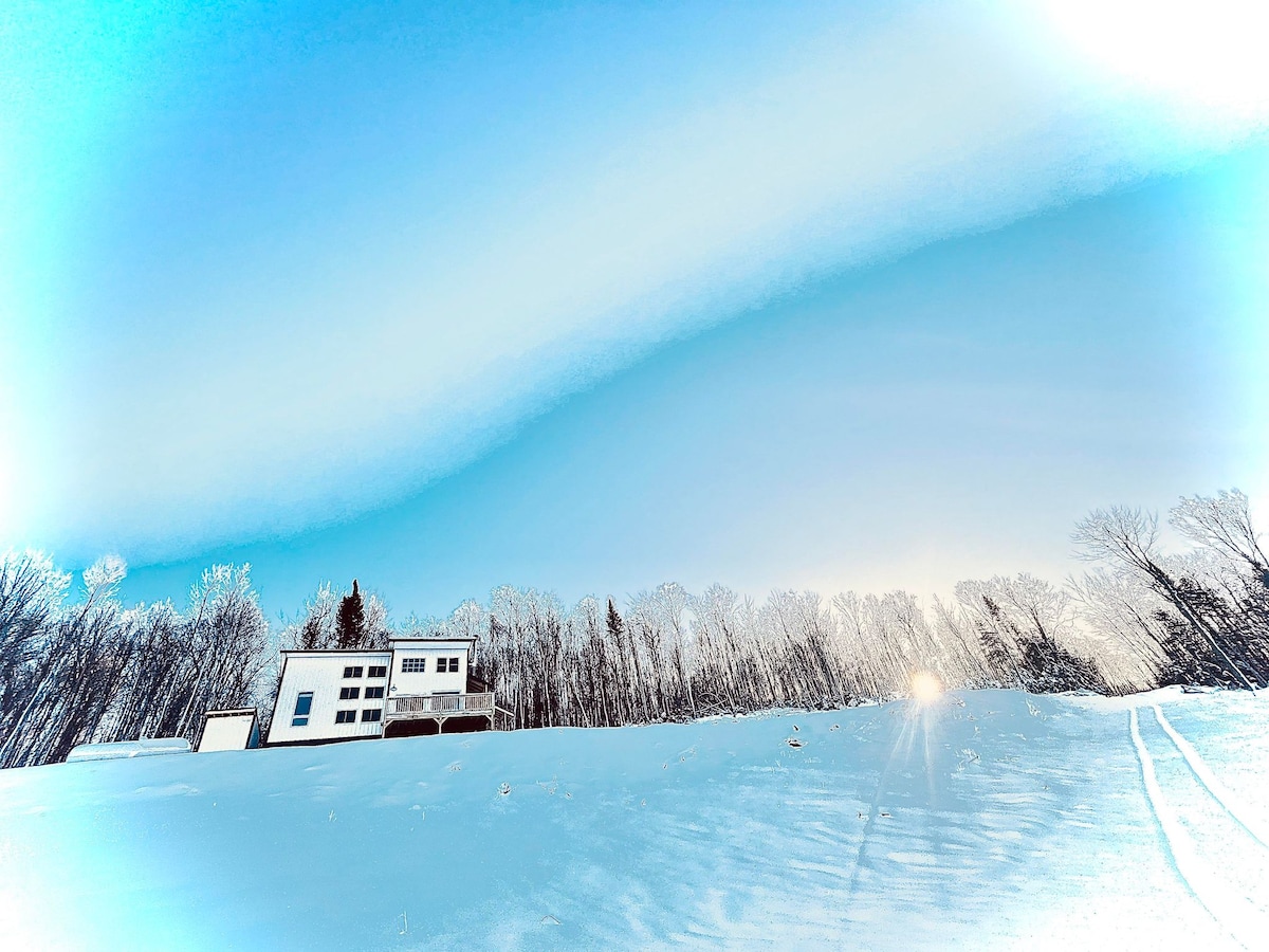 A modern cabin is set against a winter landscape, surrounded by snow-covered ground and trees. The bright blue sky contrasts with the white snow, providing a serene backdrop. Sunlight filters through the trees, illuminating the scene with a soft glow.