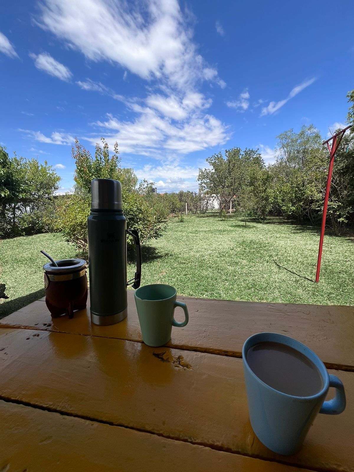 A view from a yellow table features a thermos and two mugs, one in light blue and another with a dark beverage. Lush greenery is visible in the background, with trees and a clear blue sky dotted with clouds creating a serene outdoor setting.