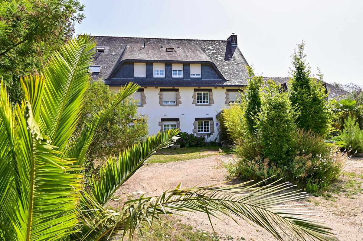 A charming building is surrounded by lush greenery and various shrubs. Large windows reflect natural light, while a mix of trees and palm foliage frame the entrance. A gravel pathway leads up to the structure, creating an inviting approach.