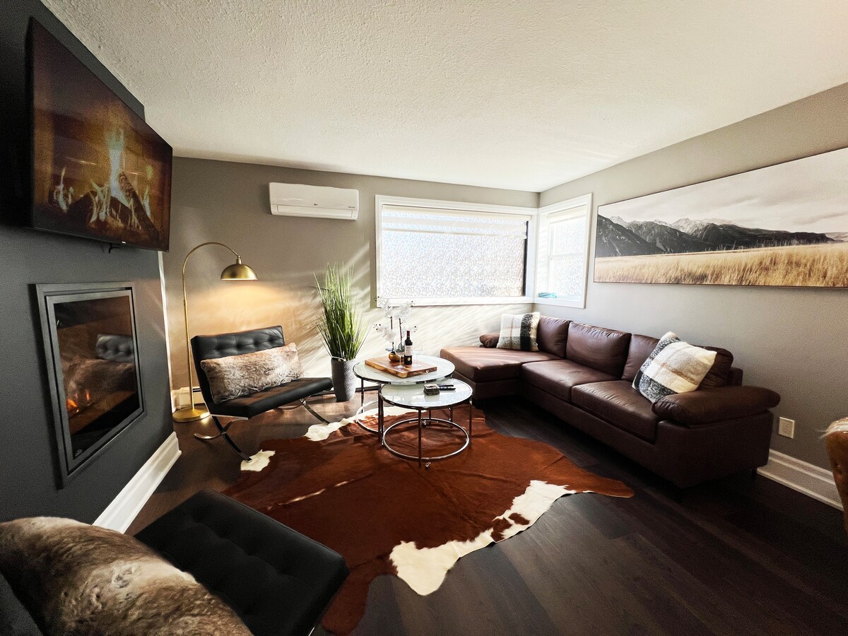 A cozy living area features a leather sectional sofa with plaid accents, complemented by a round coffee table at its center. A wall-mounted television hangs above a modern fireplace, while large windows allow natural light to brighten the space, enhanced by understated decorative elements.