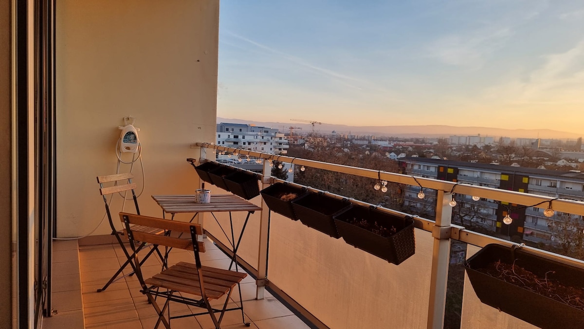 A balcony is featured, furnished with a small wooden table and two folding chairs. Flower boxes line the railing, and a scenic view of the surrounding area is visible in the background, showcasing a sunset sky filled with soft hues.