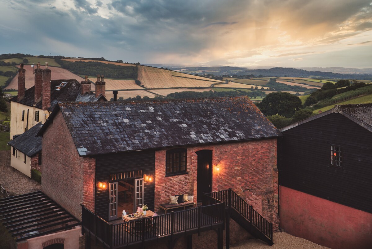 The exterior of a restored barn is shown, featuring a deck with outdoor furniture. Warm lights illuminate the building against a cloudy sky, with panoramic views of rolling countryside and fields in the background.