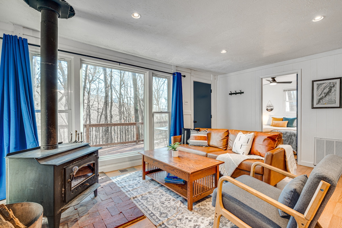 A cozy living room features a wood-burning stove and ample natural light from large windows overlooking the wooded area. Comfortable seating includes a leather sofa and wooden armchairs, positioned around a wooden coffee table, with a decorative area rug enhancing the inviting space.