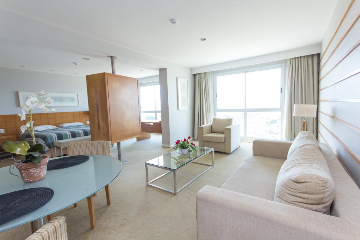 A well-lit living area showcases a comfortable sofa and a chair, complemented by a glass coffee table. A dining table with chairs is present near a large window, which offers a clear view of the surroundings. The space is finished with neutral tones and light curtains.