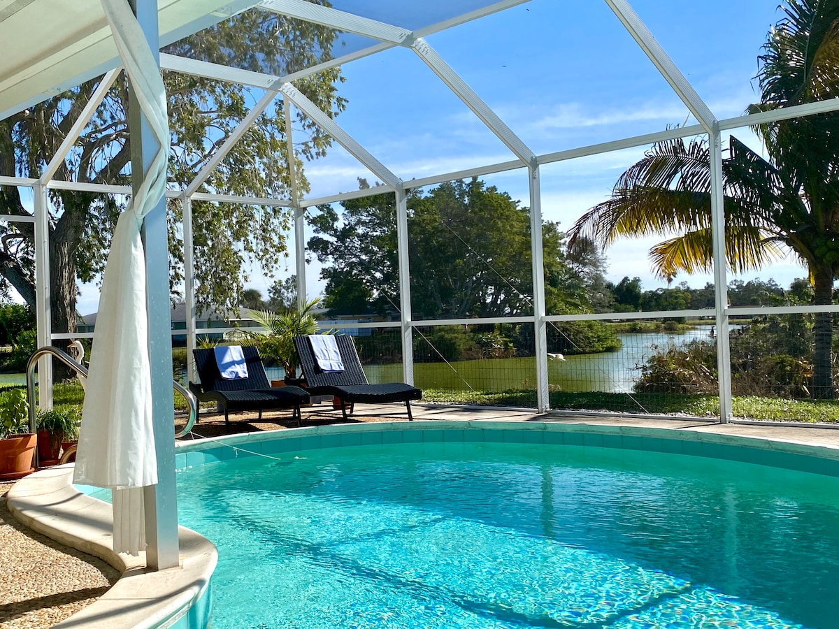 A private screened pool area is visible, featuring a clear swimming pool and two lounge chairs adorned with white towels. Lush greenery surrounds the space, with a serene lake and trees providing a natural backdrop against a clear blue sky.