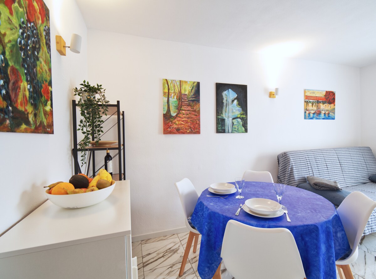 A light-filled living space features a round dining table set with plates and glasses, surrounded by modern white chairs. Colorful artwork adorns the walls, while a comfortable sofa is positioned beside a stylish shelf with decorative items. A bowl of fresh fruit adds a vibrant touch.