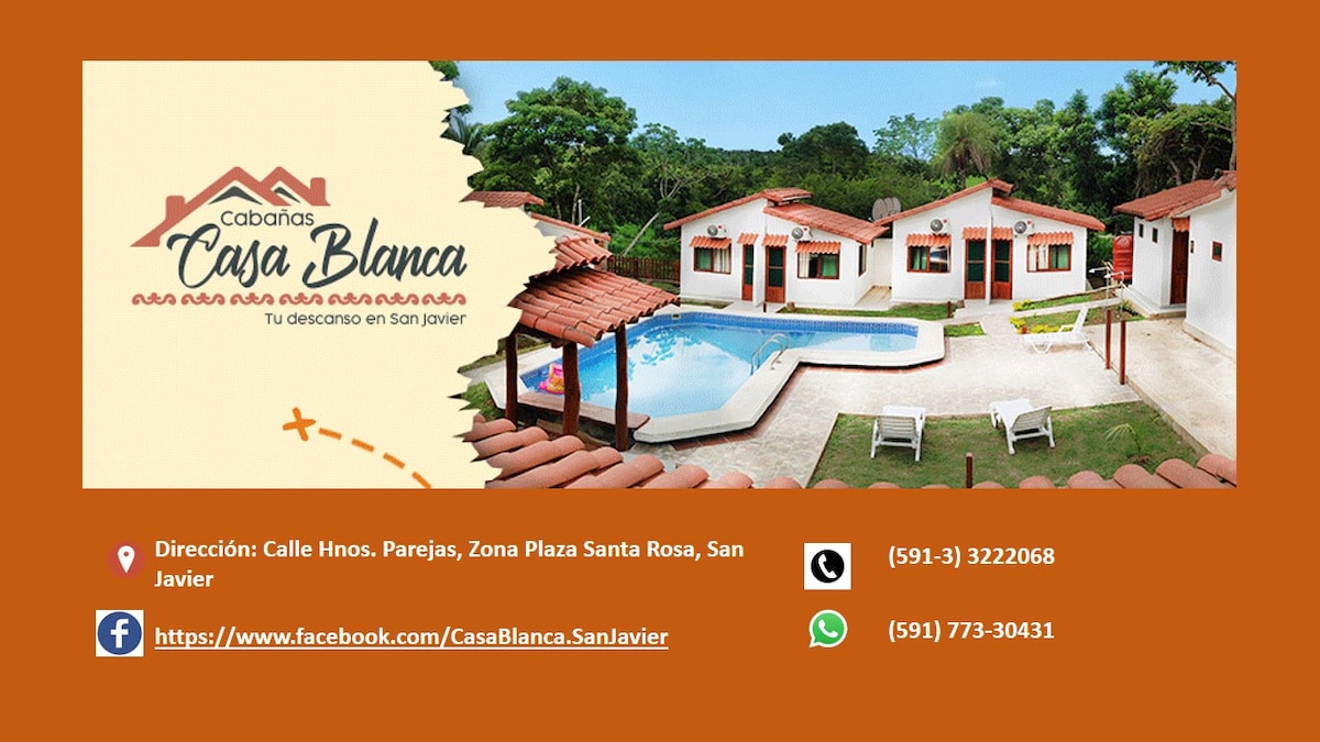 The image showcases Cabañas Casa Blanca, featuring a cluster of charming buildings with red-roofed accents. A clear pool and well-maintained gardens can be seen, providing a welcoming space for relaxation. The surrounding area is adorned with pathways and greenery, enhancing the inviting atmosphere.