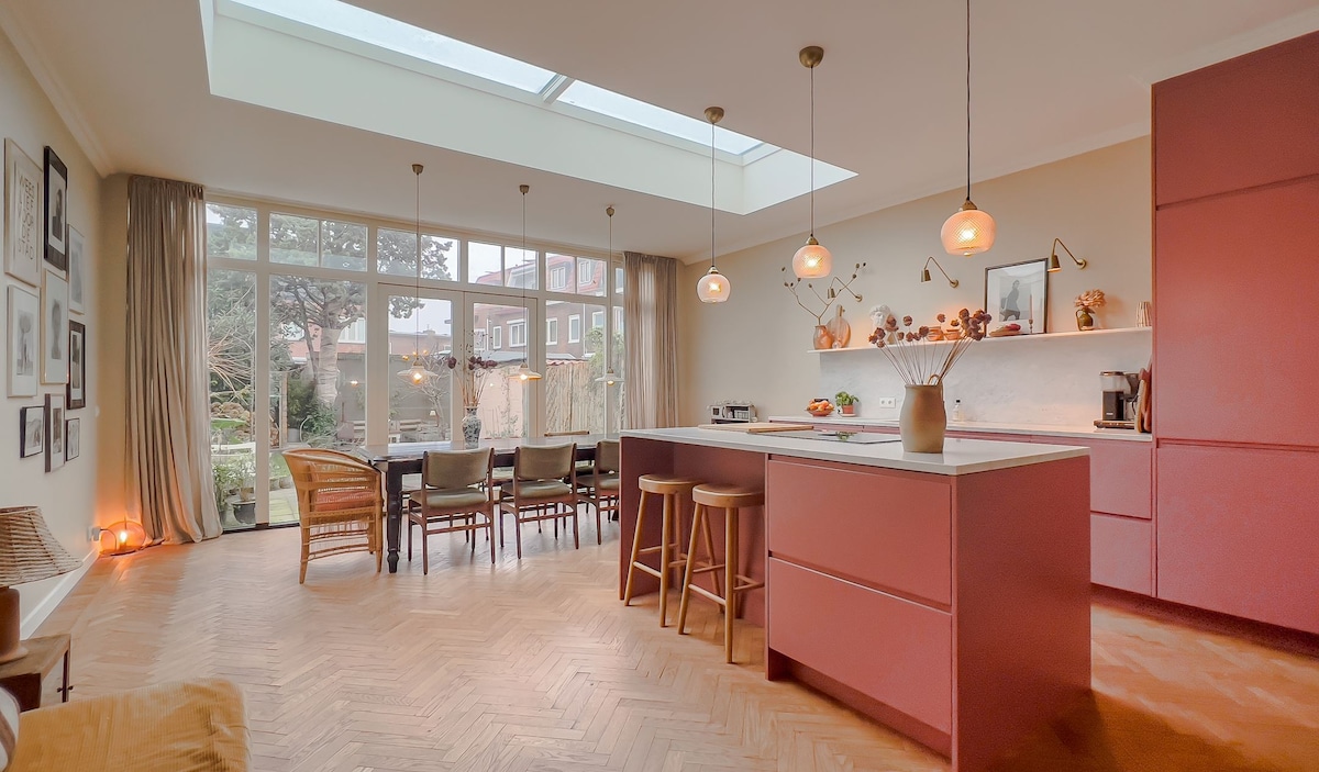 A modern kitchen and dining area features an open layout with a large skylight providing natural light. Smooth cabinetry in soft tones contrasts with the warmth of parquet flooring. A spacious dining table is surrounded by chairs, and a cozy seating area faces glass doors leading to the garden.