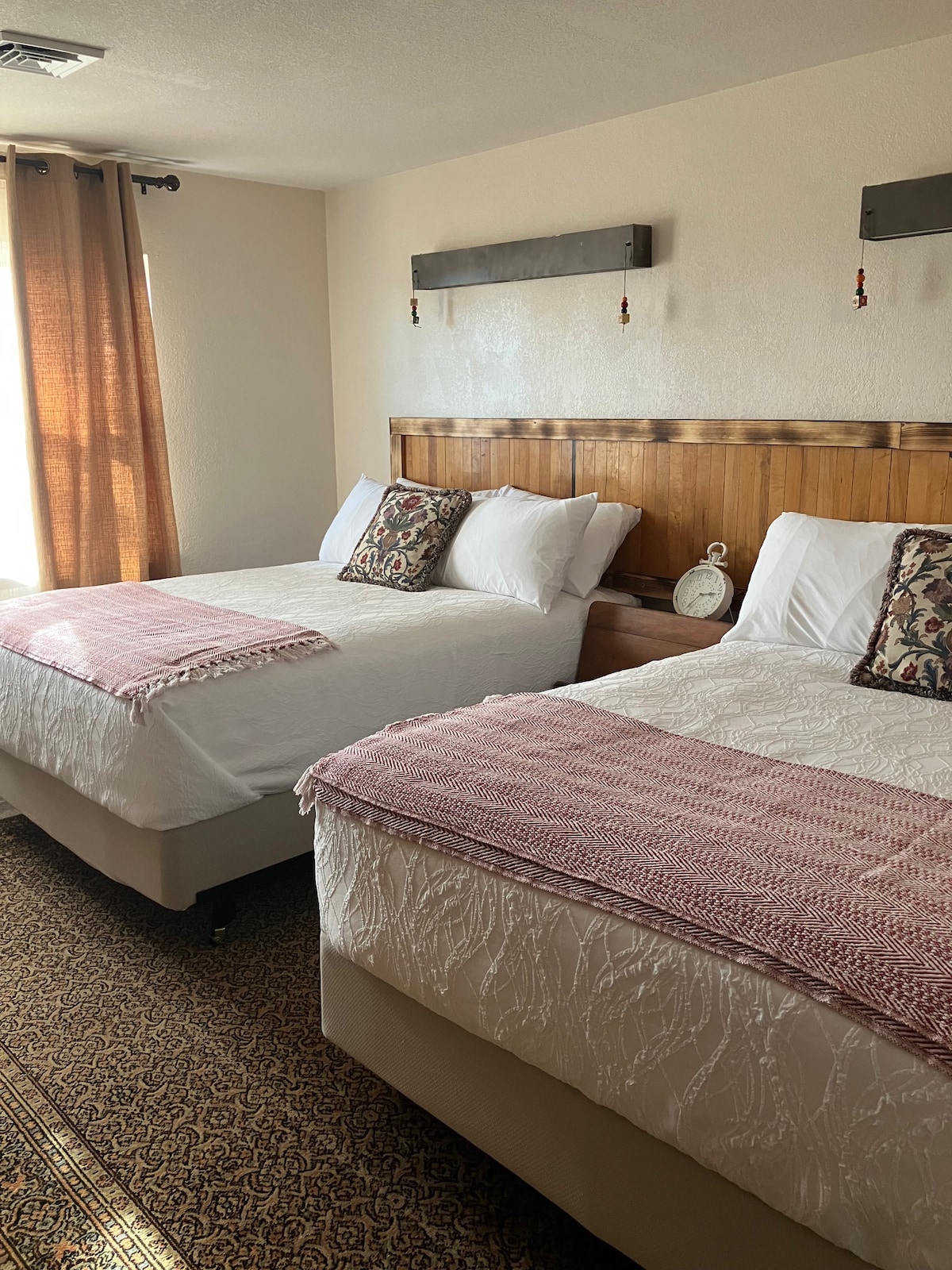 A well-furnished bedroom features two spacious beds, each dressed in crisp white linens. Decorative throw blankets and patterned pillows add subtle color. Natural light filters through the brown curtains, illuminating the wooden headboard and soft area rug beneath.