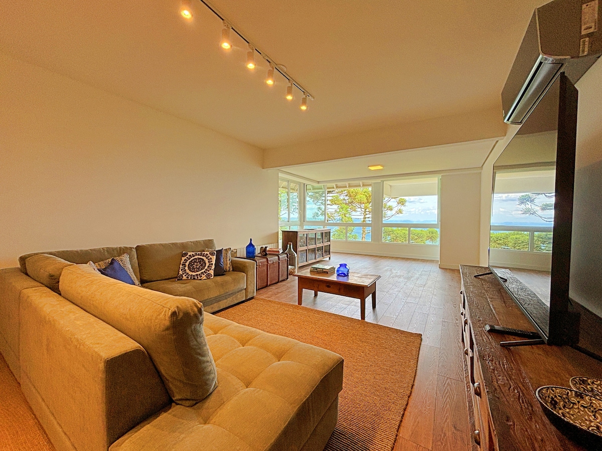 A spacious living area offers panoramic views through large windows. A comfortable sectional sofa and a wooden coffee table create a welcoming seating arrangement. Natural light fills the room, highlighting the wooden flooring and neutral decor.