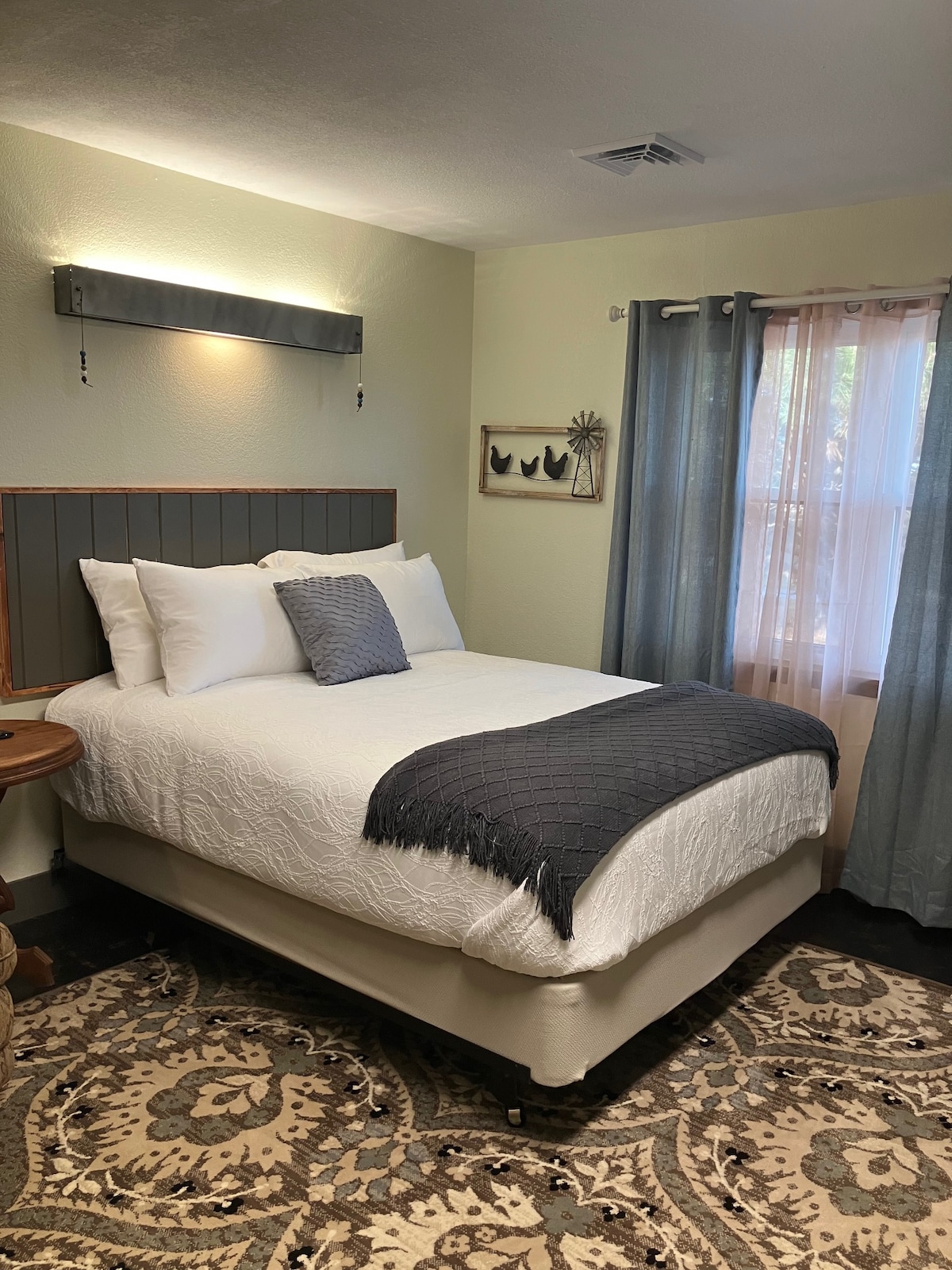 A sleeping area features a spacious bed with white linens and a dark textured throw. Natural light filters through a nearby window, adorned with light blue curtains. A circular rug with intricate patterns is positioned under the bed, complementing the warm wall colors.