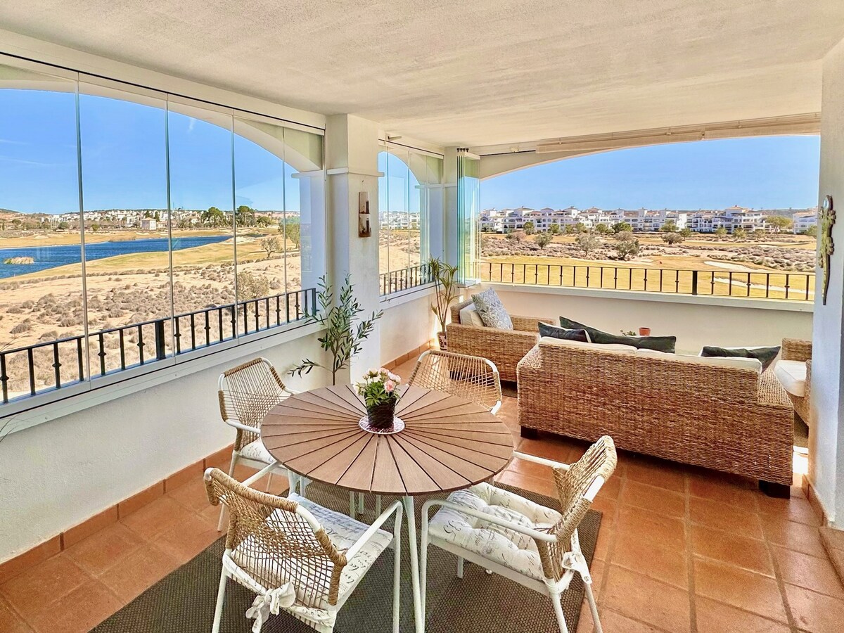 A large terrace features a round dining table surrounded by four wicker chairs. A comfortable seating area offers a spacious sofa. Glass curtains provide an unobstructed view of the lake and golf course, allowing natural light to fill the space.