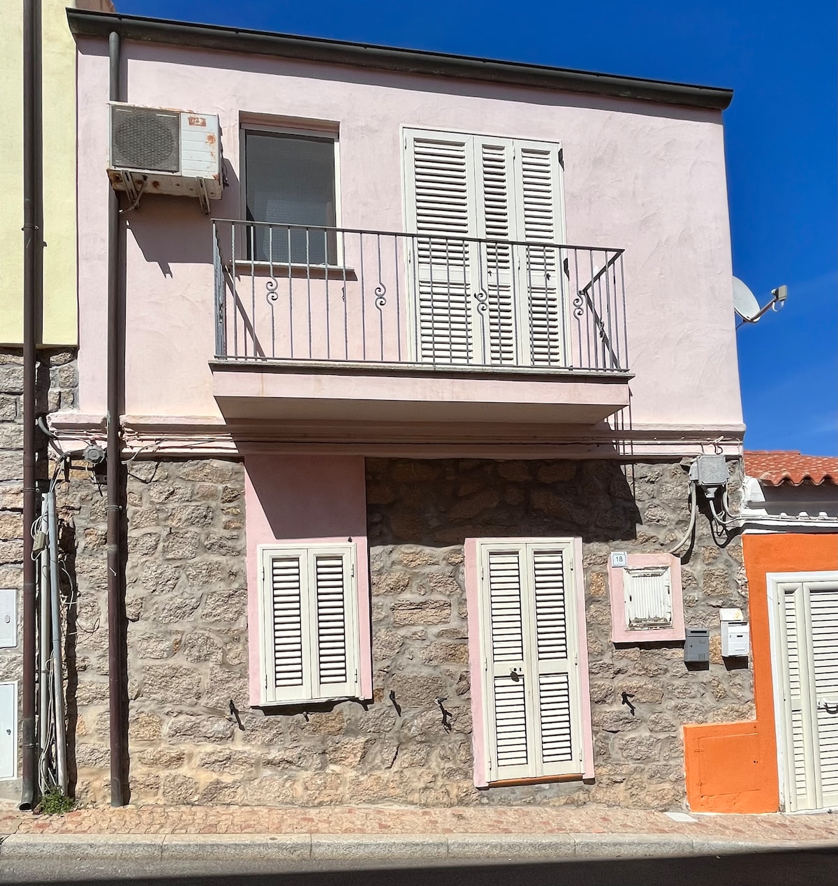 A two-story building features a charming combination of stone and pastel-colored walls. The upper floor has a balcony with a metal railing and dual shuttered windows, while the ground floor showcases additional shuttered windows and a vibrant orange entryway.