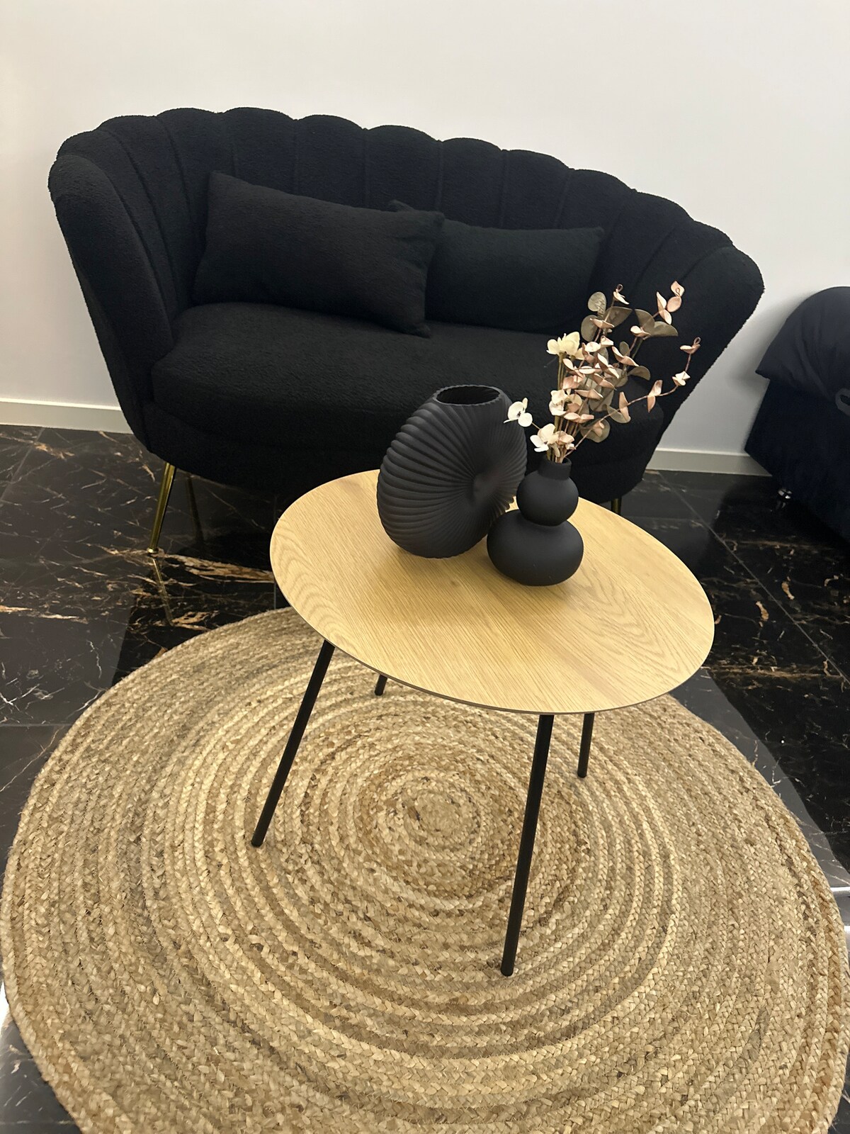 A round coffee table with slender black legs is centered on a textured rug. Two vases, one with a wavy design and another with a smooth finish, are placed atop the table. A cozy chair with a curved backrest and dark cushions is positioned nearby.