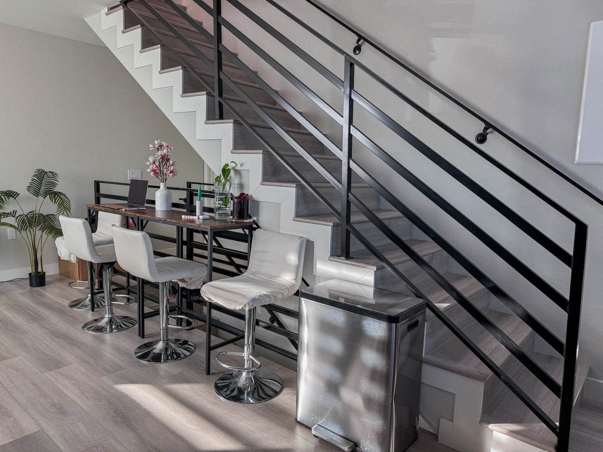 A modern dining area is located beside the staircase, featuring a sleek black table surrounded by four white bar stools. A potted plant is positioned nearby, enhancing the contemporary aesthetic of the space. Light filters through the area, creating a bright atmosphere.