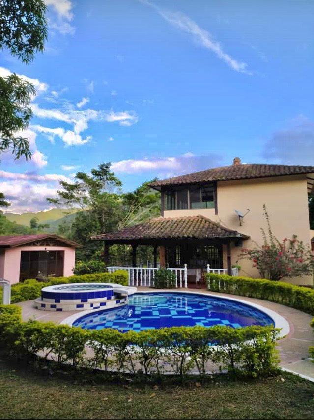 A two-story house is surrounded by vibrant greenery and features a circular pool with a decorative tile pattern. A covered seating area is located next to the pool, offering an inviting outdoor space. The sky is clear with soft clouds, creating a serene backdrop.