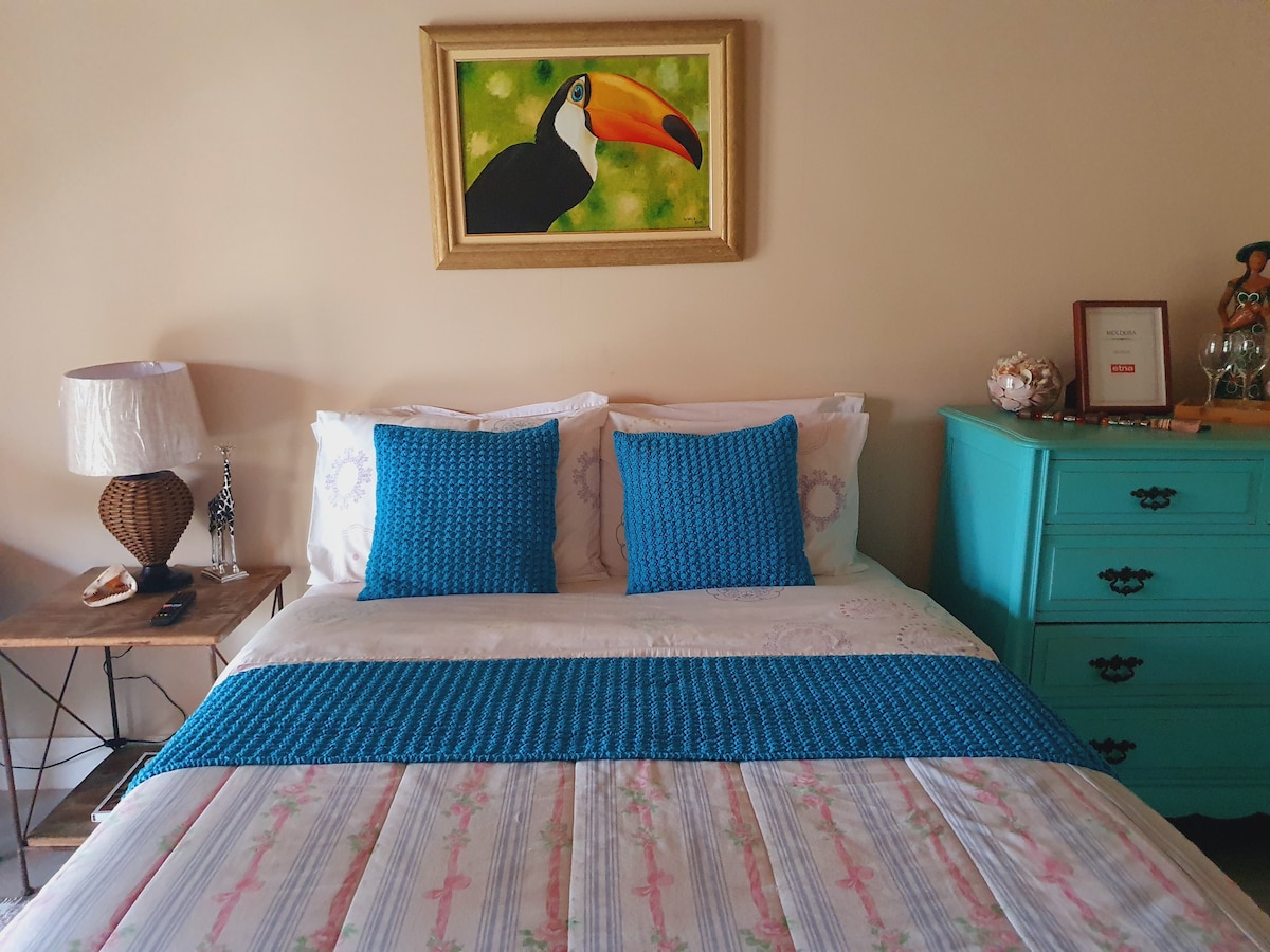 A tranquil bedroom features a spacious bed adorned with a patterned quilt. Two bright blue pillows add a pop of color, and a painting of a toucan hangs above the bed. A bedside lamp and a teal dresser complete the serene space.